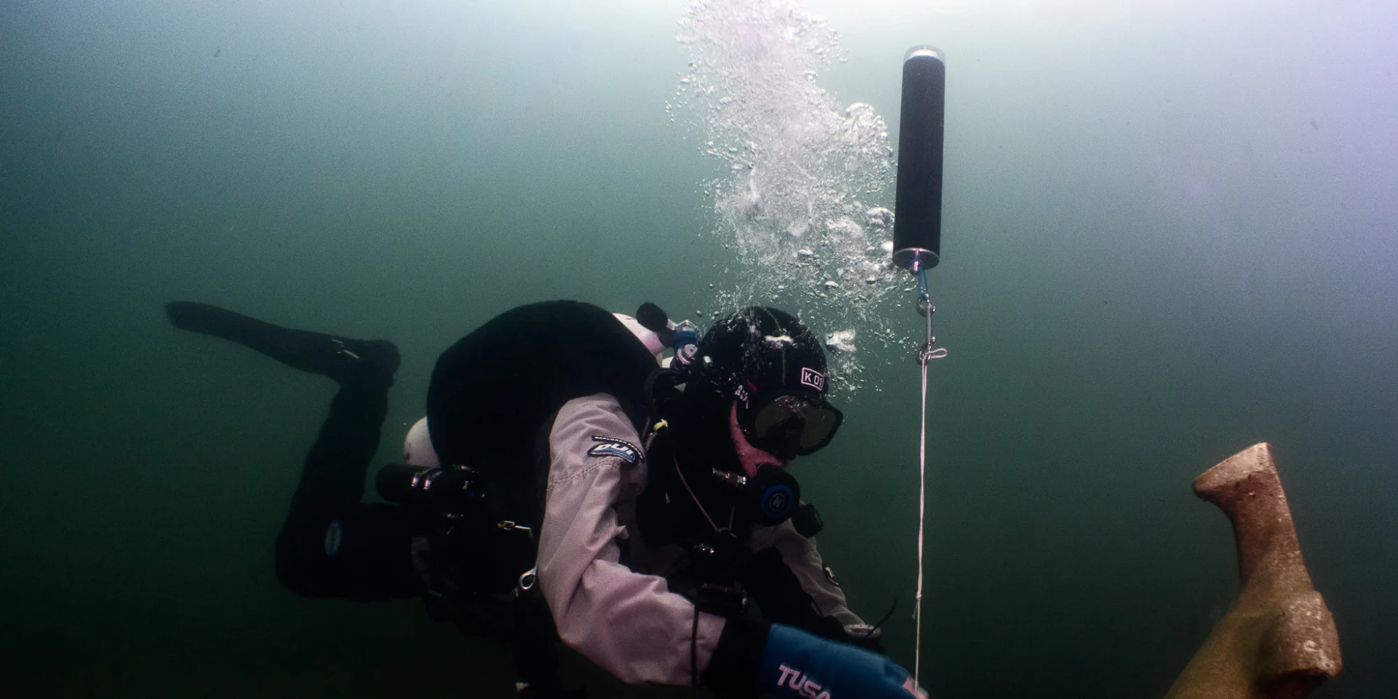 A diver on the bottom with his torch lit, the buoy above him on the line, bubbles rising
